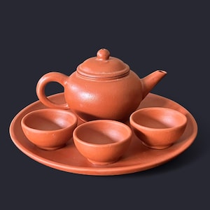 May include: A terracotta tea set, including a teapot with a lid, three small teacups, and a serving tray. The set is a warm, earthy orange-brown color. The teapot has a curved handle and spout.