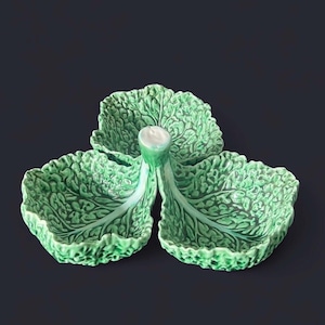 Puede incluir: Una fuente de cerámica verde con forma de col. Tiene tres secciones con forma de hoja, con vetas detalladas y una superficie texturizada. El tallo central es blanco y verde, añadiendo realismo.