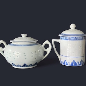 May include: White porcelain tea set with blue floral and geometric patterns. The set includes a lidded teapot with two handles and a lidded mug with a handle. The teapot has small holes and the mug has a blue border.