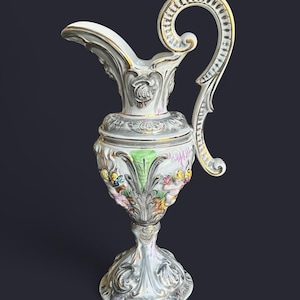Puede incluir: Jarra de porcelana ornamentada, con un asa alta y curvada y un pico. La jarra es principalmente blanca con detalles plateados y dorados, y presenta diseños florales y de hojas coloridas. La base es de estilo pedestal.