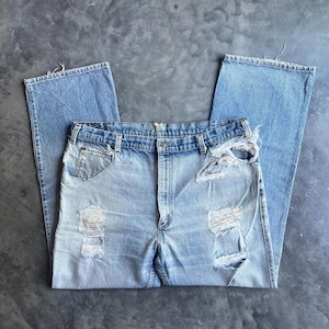 May include: Light blue denim jeans featuring a straight-leg cut, a classic five-pocket design, and a button-fly closure. The fabric displays a faded wash with intentional distressing, including frayed holes on the thighs and the front pocket area.