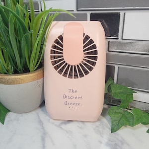 May include: A small, pink, handheld fan with a clip on the back. The fan has a white label that says "The Discreet Breeze".