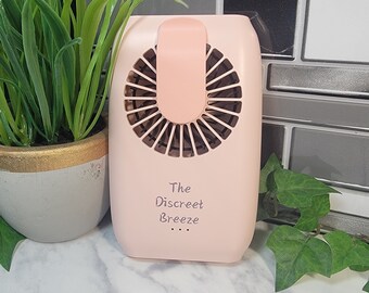 May include: A small, pink, handheld fan with a clip on the back. The fan has a white label that says "The Discreet Breeze".