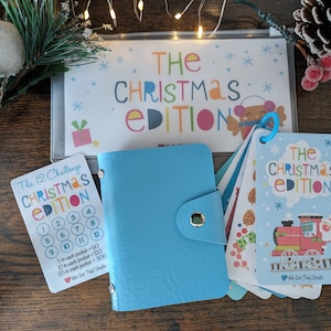 May include: A blue leather-bound booklet with a Christmas-themed 12-day challenge card. The card features a colourful design with numbers 1-12 and instructions for completing the challenge. The booklet has a zipper closure and a small metal ring.