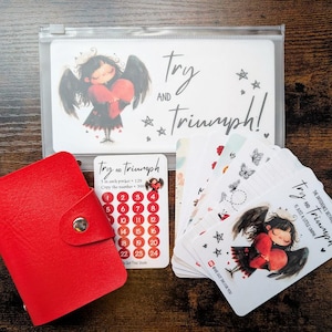 May include: A red leather card holder with a snap closure beside a set of cards. The cards feature an illustration of a girl with angel wings and the text "Try and Triumph!" on a clear plastic pouch.