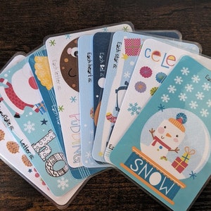 May include: A set of eight blue and white cards with winter-themed illustrations and text. Each card features a different image, such as a snowman, a snowflake, a star, a bell, a heart, a letter, a pudding, and a Santa Claus. The text on each card reads "Each snowflake is", "Each star is", "Each bell is", "Each heart is", "Each letter is", "Each star is", "Pudding", and "Snowflake is".