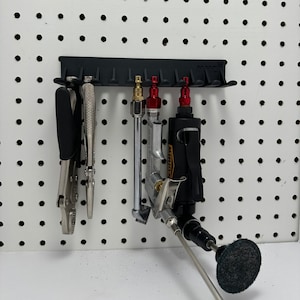 May include: A black pegboard organizer with various tools. The tools include pliers, air tools, and a rotary tool with a black abrasive pad. The tools are arranged on a white pegboard with evenly spaced holes.