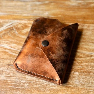 Puede incluir: Una cartera de cuero artesanal con un acabado de cobre envejecido único. La cartera tiene un cierre de solapa asegurado por un broche negro. Los bordes están cuidadosamente cosidos con hilo negro. Está diseñada para guardar tarjetas y efectivo.