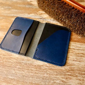 May include: A bi-fold card holder made of smooth navy blue leather with visible dark stitching. The interior features a thumb cutout on the left pocket and a diagonal cut on the right pocket, revealing a black inner lining and a grey suede texture.