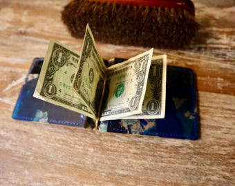 May include: A blue and white patterned wallet with several one-dollar bills fanned open. The wallet is open, displaying the bills, and is set against a wooden surface. A brown object is in the background.