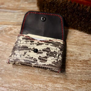 May include: Open, small, patterned wallet with a black interior and red stitching. The exterior features a reptile-like pattern in shades of brown and cream. A black snap closure is visible.