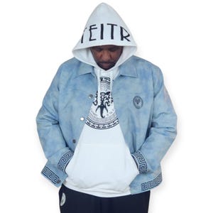 Pu&ograve; includere: Una giacca di jeans blu chiaro con una felpa bianca sotto. La giacca ha un motivo tie-dye e disegni a chiave greca sui polsini e sul collo. La felpa ha una grafica in bianco e nero di una creatura mitologica con il testo "EITR" sul cappuccio.