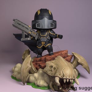 Può includere: Una statuetta di un personaggio in armatura nera e oro, con una pistola d'argento. La figura è in piedi su una formazione rocciosa marrone sopra una base beige a forma di mostro con denti affilati. Il testo "Painting suggestion" è in basso.