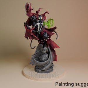 Pode incluir: Uma figura detalhada do personagem de banda desenhada Spawn, com uma capa vermelha e correntes. A figura está posada dinamicamente sobre uma base esculpida com um anjo. Um efeito de energia verde também está presente. A base tem o texto "Remerinds" e "Painting suggestion".