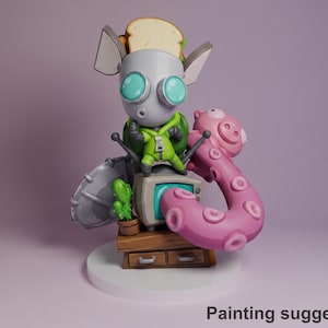 Op de afbeelding: Een grillig beeldje met een grijs, buitenaards personage met grote turquoise ogen, een groene jas en een sandwich op zijn hoofd. Een roze octopus-achtig aanhangsel en een retro tv maken de scène compleet.