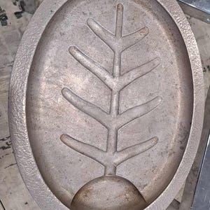 May include: A silver oval-shaped tray with a raised design of a stylised tree branch in the centre. The tray has a textured surface and a small indentation on the bottom.