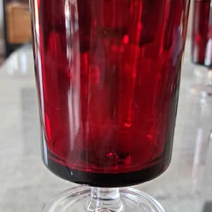 Könnte beinhalten: Ein rubinrotes Weinglas mit klarem Stiel und Fuß. Das Glas hat eine zylindrische Schale und einen schwarzen Rand. Es steht auf einer reflektierenden Oberfläche. Die Beleuchtung hebt die Farbe und Form des Glases hervor.