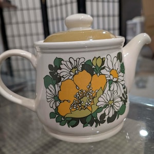 以下が含まれることがあります： クリーム色のセラミックティーポット。黄色と白の花と緑の葉のフローラルデザイン。蓋はタン色で、ハンドルと注ぎ口は湾曲しています。キッチン用品。