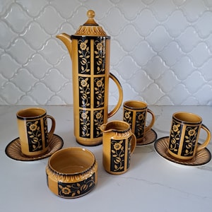 Może przedstawiać: Zestaw ceramicznych filiżanek do herbaty i czajnika z brązowym i czarnym wzorem kwiatowym. Czajnik jest wysoki i cylindryczny z pokrywką i uchwytem. Filiżanki do herbaty są mniejsze i zaokrąglone z uchwytem. Zestaw jest ozdobiony kwiatowym wzorem w kolorze czarnym i brązowym.