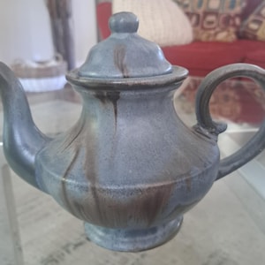 Puede incluir: Tetera de cerámica en un tono azul grisáceo apagado con vetas marrones. Cuenta con tapa, boquilla y asa curvada. La tetera se apoya sobre una pequeña base. Medidas en centímetros.