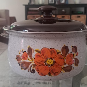 Puede incluir: Una olla vintage de esmalte moteado con tapa y asas de color marrón oscuro. La olla presenta un diseño floral en naranja, marrón y amarillo. Esta olla es adecuada para su uso en la estufa.