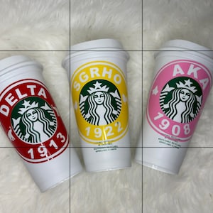 May include: Three white reusable cups with black lids. Each cup has a different coloured background and a green Starbucks logo with a woman's face in the centre. The cups are labelled with the text "DELTA 1913", "SGRHO 1922", and "AKA 1908".