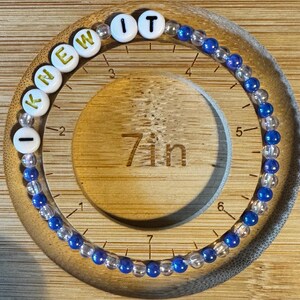 May include: A circular beaded bracelet featuring alternating clear and blue beads, with white letter beads spelling out I KNEW IT. The bracelet is positioned on a wooden measuring tool that clearly indicates a circumference of 7 inches.