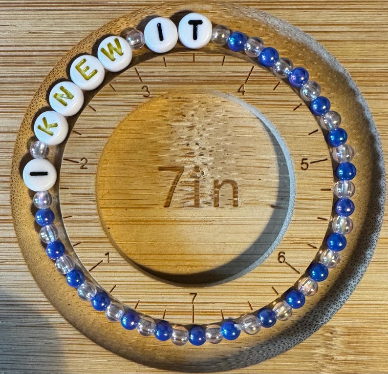 May include: A circular beaded bracelet featuring alternating clear and blue beads, with white letter beads spelling out I KNEW IT. The bracelet is positioned on a wooden measuring tool that clearly indicates a circumference of 7 inches.