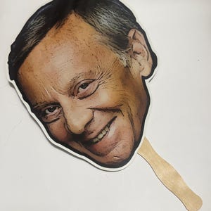 May include: A novelty hand fan featuring a smiling face with a wooden handle. The face is a printed image of a man with dark hair and a tan complexion. The fan is white around the edges.