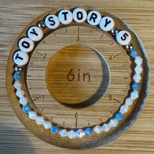 May include: A circular bracelet featuring white letter beads spelling TOY STORY 5, interspersed with small silver-toned metallic beads and faceted light blue and iridescent white beads, positioned on a wooden measuring tool showing a 6-inch diameter.