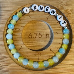 May include: A circular beaded bracelet featuring alternating light blue and yellow translucent beads, interspersed with small silver-toned spacer beads. White letter beads spell out the phrase I KNEW IT. A ruler indicates a 6.75 inch diameter.