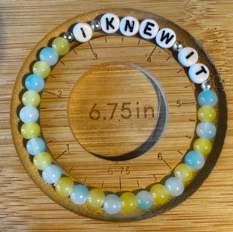 May include: A circular beaded bracelet featuring alternating light blue and yellow translucent beads, interspersed with small silver-toned spacer beads. White letter beads spell out the phrase I KNEW IT. A ruler indicates a 6.75 inch diameter.