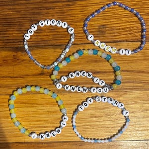 May include: Five beaded bracelets featuring white letter beads and colorful glass beads. Two bracelets display the text TOY STORY 5, two display the text I KNEW IT, and one displays the text I KNEW YOU. The beads are arranged in circular patterns.