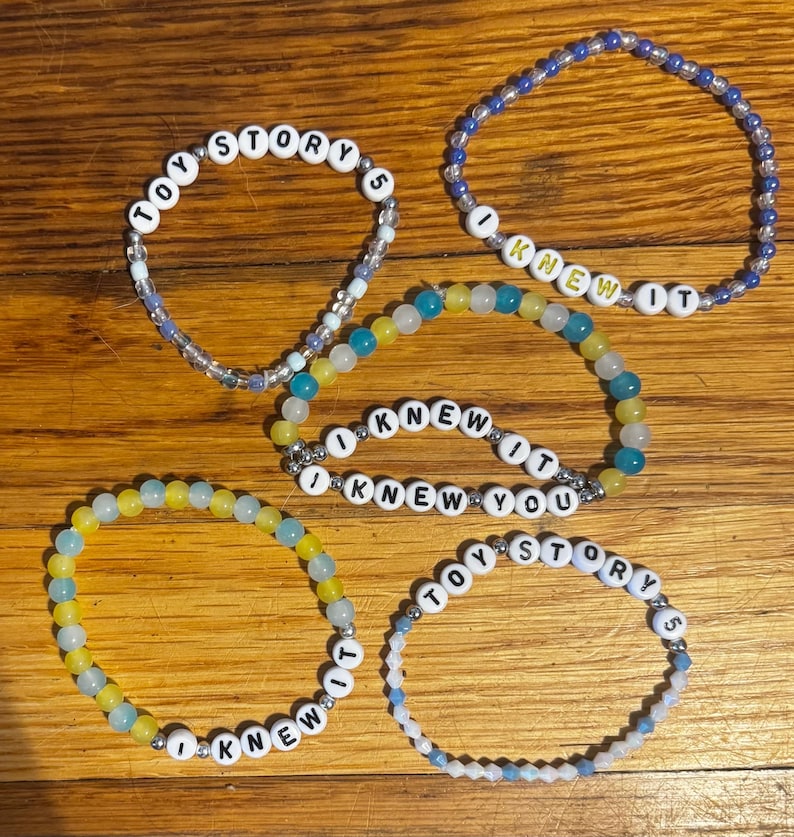 May include: Five beaded bracelets featuring white letter beads and colorful glass beads. Two bracelets display the text TOY STORY 5, two display the text I KNEW IT, and one displays the text I KNEW YOU. The beads are arranged in circular patterns.