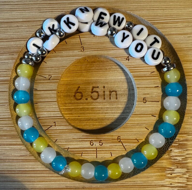 May include: A circular beaded bracelet featuring white letter beads spelling I KNEW YOU, interspersed with metallic silver beads and alternating yellow, white, and blue translucent beads. The bracelet is placed on a 6.5 inch measuring tool.