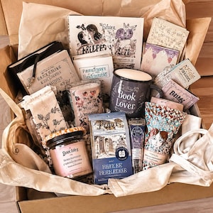 May include: A gift box containing a dark grey mug with the text Book Lover, a candle in a glass jar labelled Bookiez, several books with illustrated covers, a small tube of cream, a bag of coffee, and a white fabric pouch nestled in brown paper.