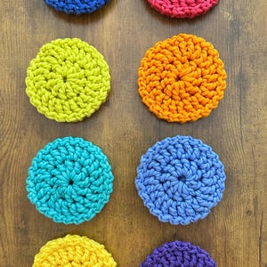 May include: Eight colorful, crocheted circular coasters in shades of blue, pink, green, orange, turquoise, purple, and yellow. The coasters are arranged on a wooden surface, showcasing their vibrant colors and textured design. Each coaster is approximately 4 inches in diameter.