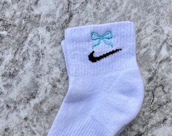 May include: White ankle socks with a black Nike swoosh logo and a light blue bow embroidered on the top. The socks have a black size tag on the heel.