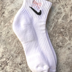 May include: White ankle socks with a black Nike swoosh logo and a pink bow embroidered on the top.
