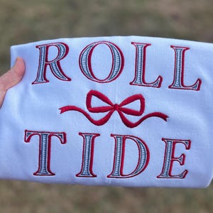 May include: A white sweatshirt featuring the embroidered text ROLL TIDE in red and grey block lettering. Between the two words is a red embroidered bow. The design is centered on the front of the garment, showcasing clean stitching.