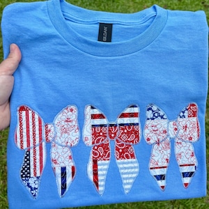May include: Pale blue t-shirt with five decorative bow appliques. The bows feature red, white, and blue patterns, including stripes, floral designs, and a bandana print. The shirt is folded and displayed on a green grassy surface.