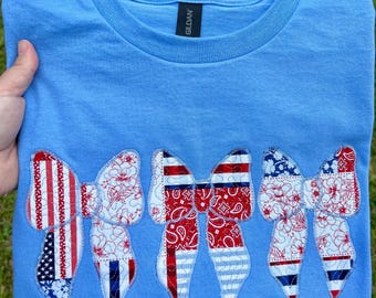 Peut inclure: T-shirt bleu clair orné de cinq nœuds décoratifs. Les nœuds présentent des motifs rouges, blancs et bleus, notamment des rayures, des motifs floraux et un imprimé bandana. Le t-shirt est plié et présenté sur une surface herbeuse verte.