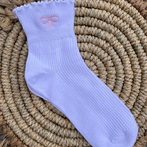 May include: White ankle socks with a pink embroidered bow on the front. The socks have a frilled top.