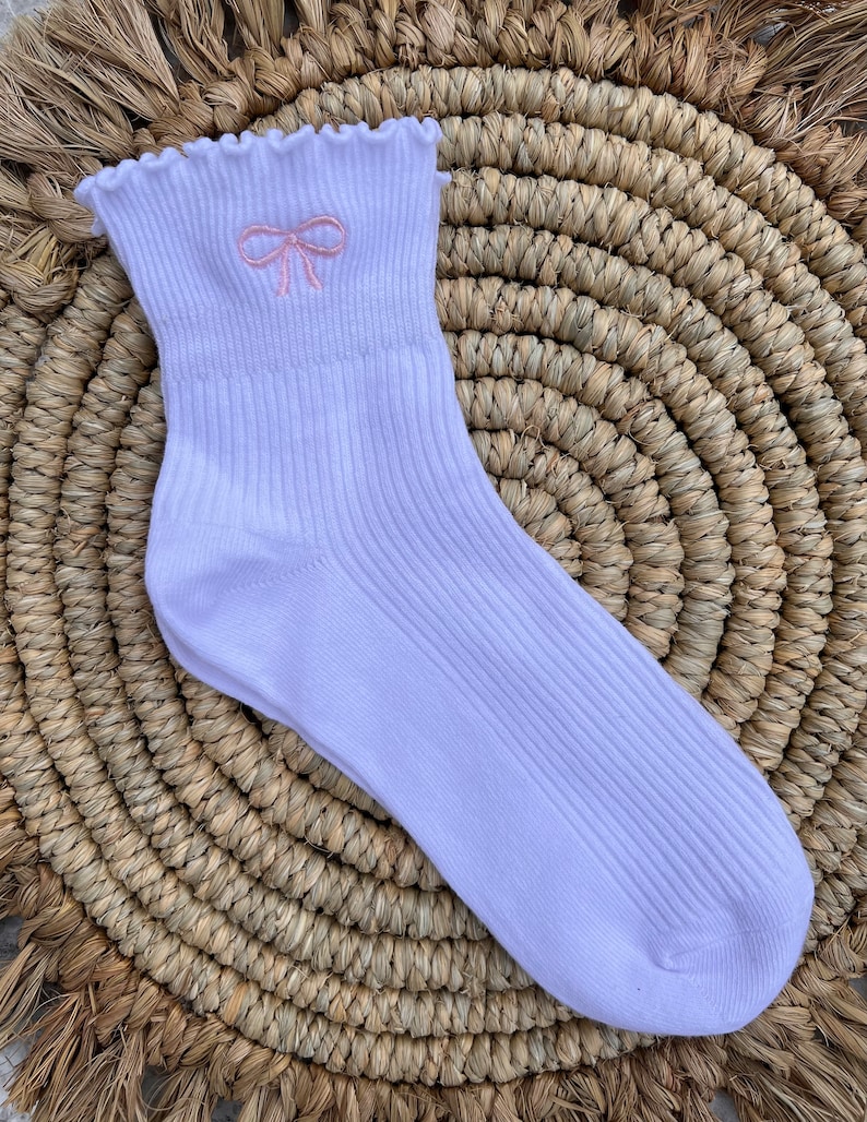 May include: White ankle socks with a pink embroidered bow on the front. The socks have a frilled top.