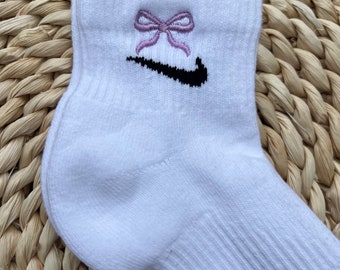 May include: White ankle socks with a black Nike swoosh logo and a purple bow embroidered on the top.