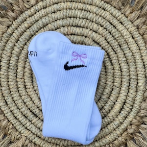 May include: White socks with a black Nike swoosh and a pink bow embroidered on the front. The socks are folded and displayed on a woven mat.