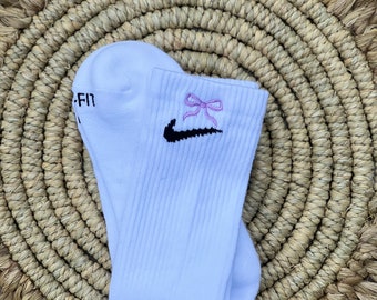 Peut inclure: Des chaussettes blanches avec un swoosh Nike noir et un nœud rose brodé sur le devant. Les chaussettes sont pliées et présentées sur un tapis tissé.