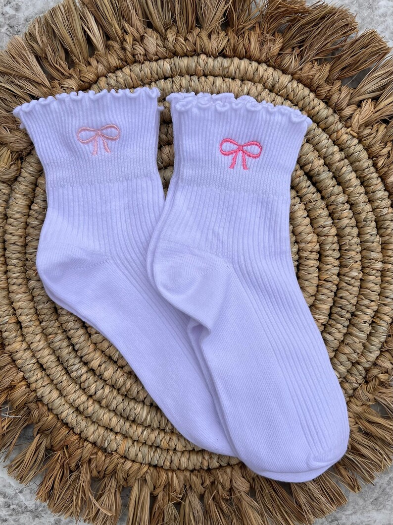 May include: A pair of white ankle socks with a pink bow embroidered on each sock. The socks have a frilled top.
