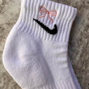 May include: White ankle socks with a black Nike swoosh logo and a pink bow embroidered on the top.