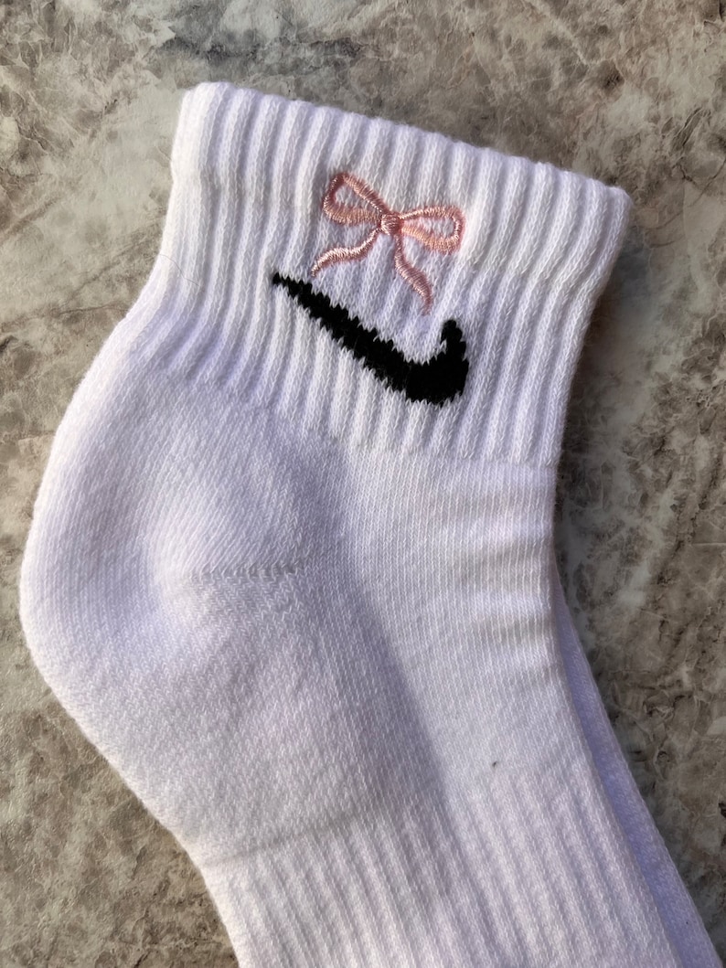 May include: White ankle socks with a black Nike swoosh logo and a pink bow embroidered on the top.